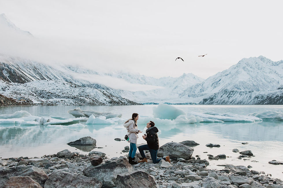 Man kneels proposing to woman by an icy lake with snow-capped mountains and birds in the sky, creating a serene, romantic scene.