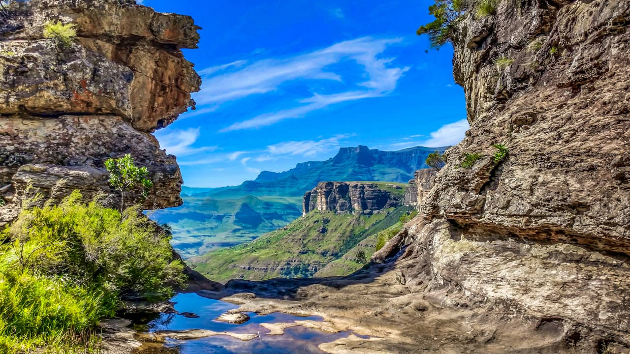 Dramatic mountain landscape with rocky cliffs framing a lush valley beneath a clear blue sky. A small reflective pool adds tranquility to the scene. South Africa, Drakensberg, Natural Regions, Touristic regions, Cape Wilderness, Nature, Wildlife, Plants, Animals.