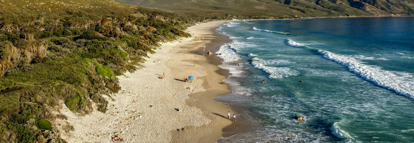 A serene beach scene with white sands, turquoise waves, and a backdrop of rugged mountains under a clear blue sky, conveying tranquility and natural beauty. South Africa, Cape Wilderness, Nature, Wildlife, Plants, Animals, Beaches.