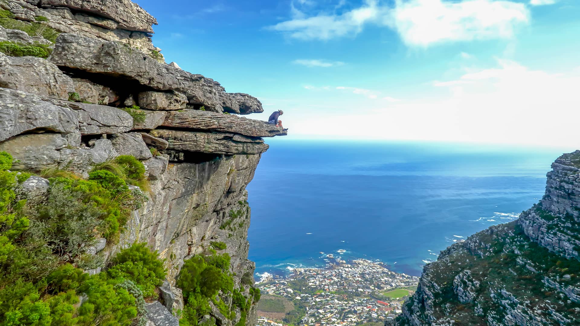 A person sits on a rocky cliff edge, overlooking a scenic ocean view under a clear blue sky. The scene conveys a sense of adventure and awe. South Africa, Cape Wilderness, Nature, Wildlife, Plants, Animals, Cape Peninsula, Cape Town.