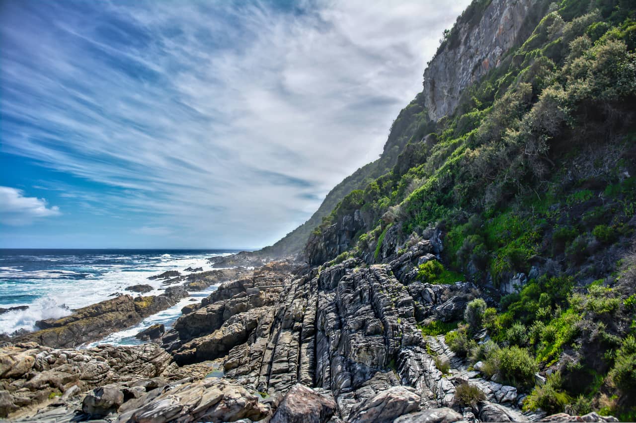 Rocky coastline with jagged cliffs covered in lush greenery. The ocean waves crash against the rocks under a vibrant blue sky with wispy clouds, creating a dramatic and serene scene. South Africa, Cape Wilderness, Natural Regions, Touristic regions, Garden Route, Nature, Wildlife, Plants, Animals.