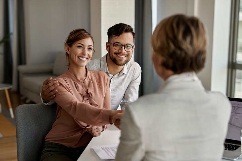 A happy couple shaking hands with a businesswoman, seen from behind, after closing a project deal.