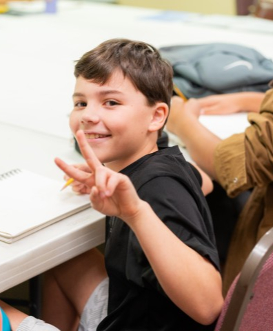 A lower elementary student give the peace sign to a photographer during a hands-on STEM learning activity