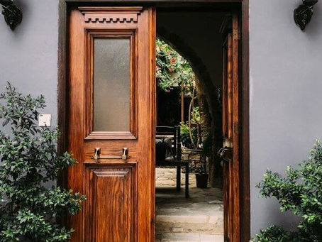 Elegant wooden front door partially open, revealing a lush garden courtyard with stone flooring and archway, framed by neatly trimmed shrubs and dark gray exterior walls.