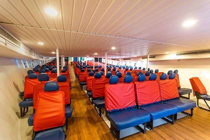 Empty ferry cabin with rows of blue seats covered in red, wood floor, and bright ceiling lights, viewed down the aisle