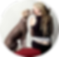A young woman with tattoos sits on a red stool, facing away from the viewer. She sports long blond dreadlocks, large earrings, and shares a moment with a Mariposa Xoloitzcuintli sitting on her left side, affectionately kissing her cheek
