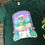 Thumbnail: Forest green tee shirt with a printed image on the front. Image is of a girl wearing a cowboy hat, sheer skirt, striped tube socks & peach roller skates strolling in the desert
