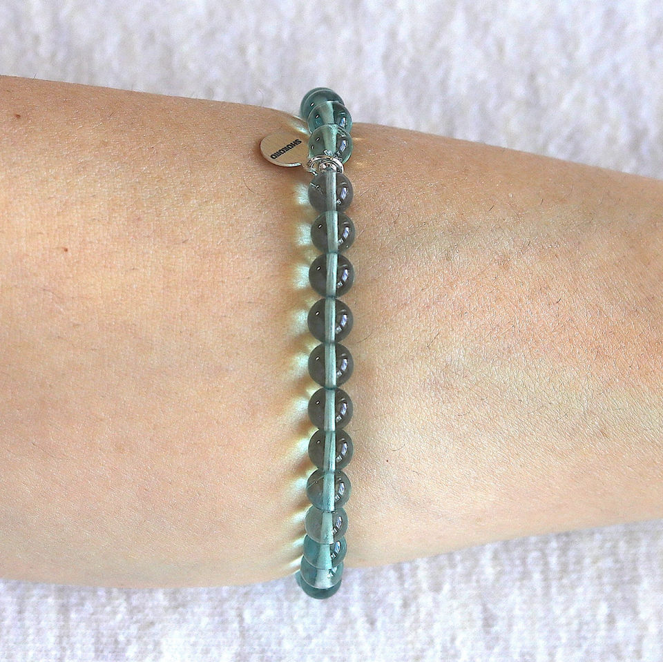 Wrist with a teal blue fluorite beaded bracelet and a small silver tag reading "shokoro". A simple, elegant design.