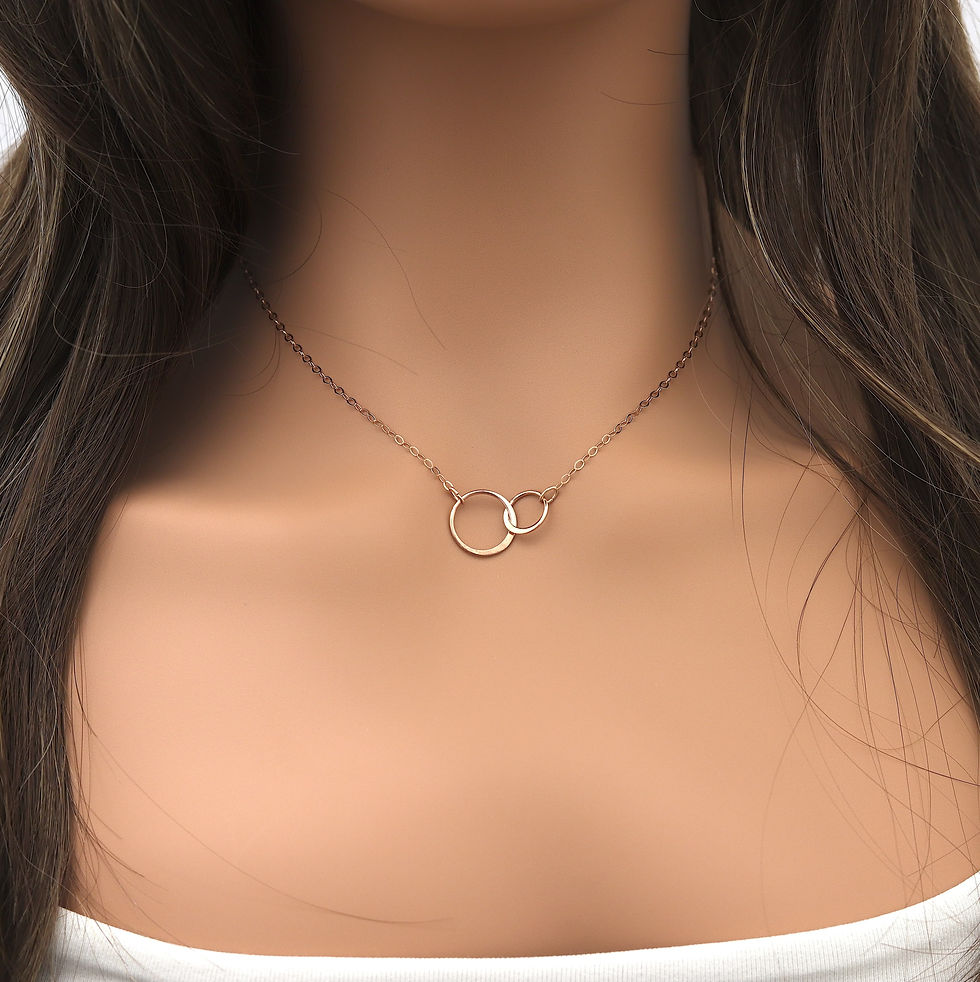 Woman wearing a rose gold double-circle necklace over a white top. Brown hair frames her neck. The mood is elegant and minima