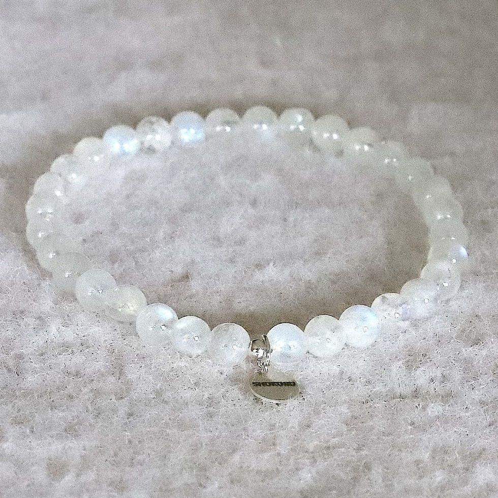 White moonstone beaded bracelet with a silver charm on a soft, textured beige surface. The charm has small, text. Minimalist