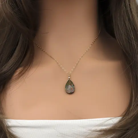 Gold chain with a teardrop unakite stone pendant on a woman's neck. She is wears a white top, creating a simple, elegant look