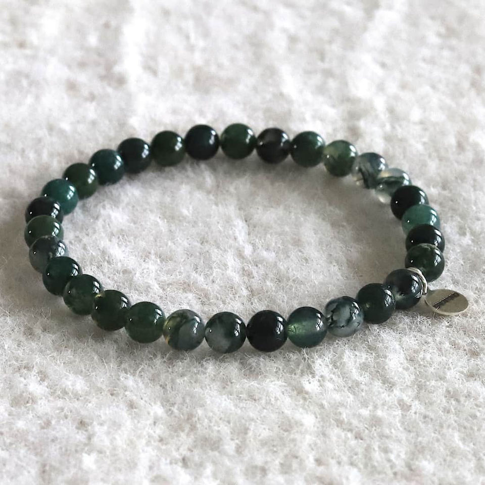 Moss agate bead bracelet on white textured fabric, featuring a small metallic charm. Mood is calm and tranquil.