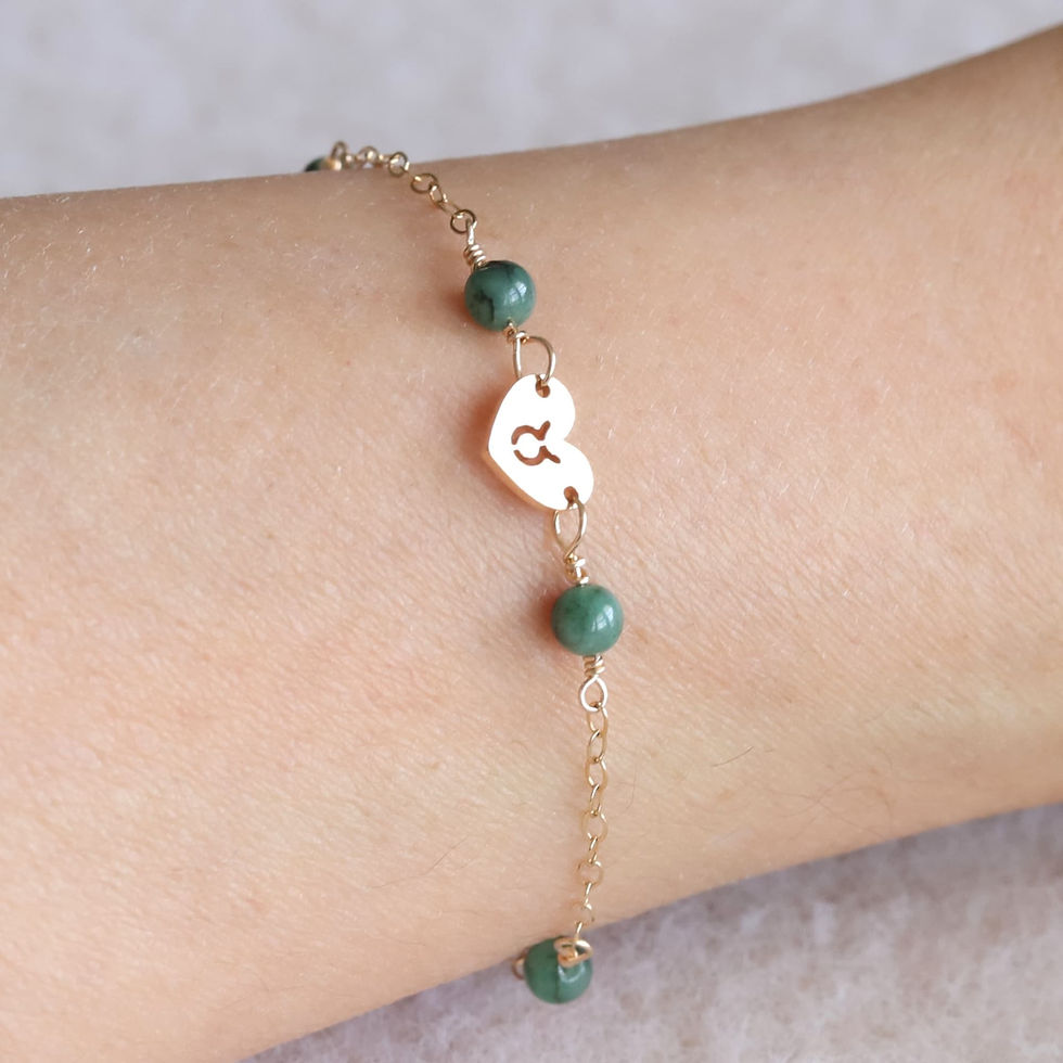 Gold chain bracelet with green beads and a heart charm on a wrist. The charm has a Taurus zodiac symbol. Light neutral mood.