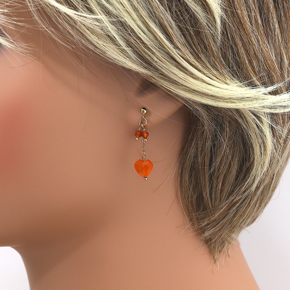 Close-up of a woman's ear with short blonde hair, showcasing an carnelian heart-shaped dangling earring. Neutral background.