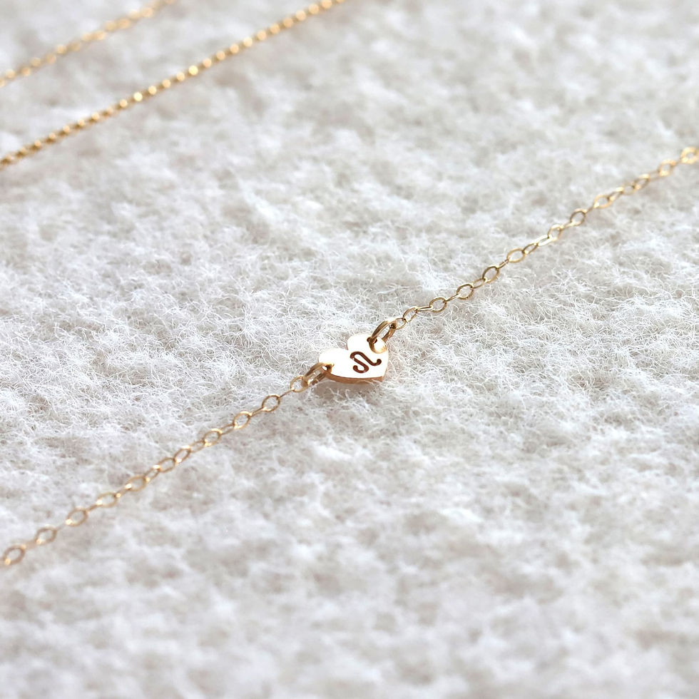 Gold chain anklet with a heart-shaped charm featuring a Leo zodiac symbol on soft white fabric. Elegant and delicate design.