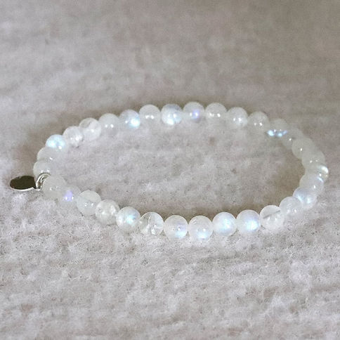 White moonstone beaded bracelet with subtle iridescence on a soft, textured beige background. A small, dark tag is attached,