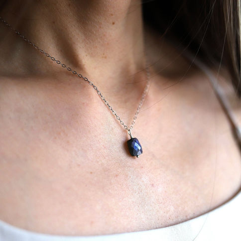 A close-up of a person wearing a silver chain necklace with a labradorite gemstone pendant. The background is blurred and neu