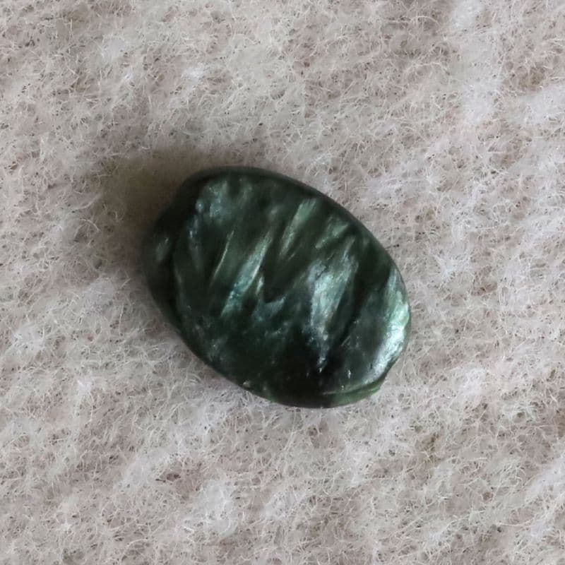 Shokoro Handmade private collection of Natural Seraphinite, dark green oval stone on pale fuzzy fabric, shown in close-up with a glossy, ridged surface.