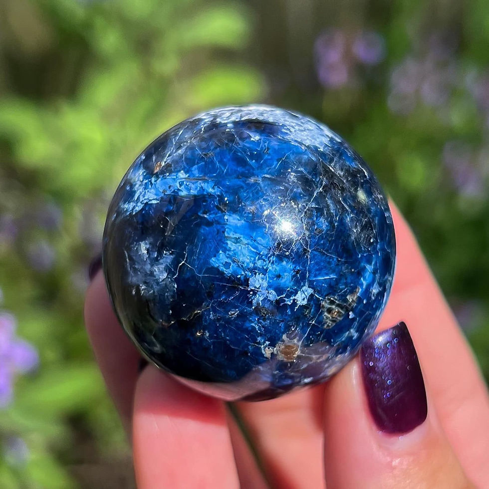 Close-up of a hand with purple nails holding a shiny blue sphere against a blurred green and purple floral background.