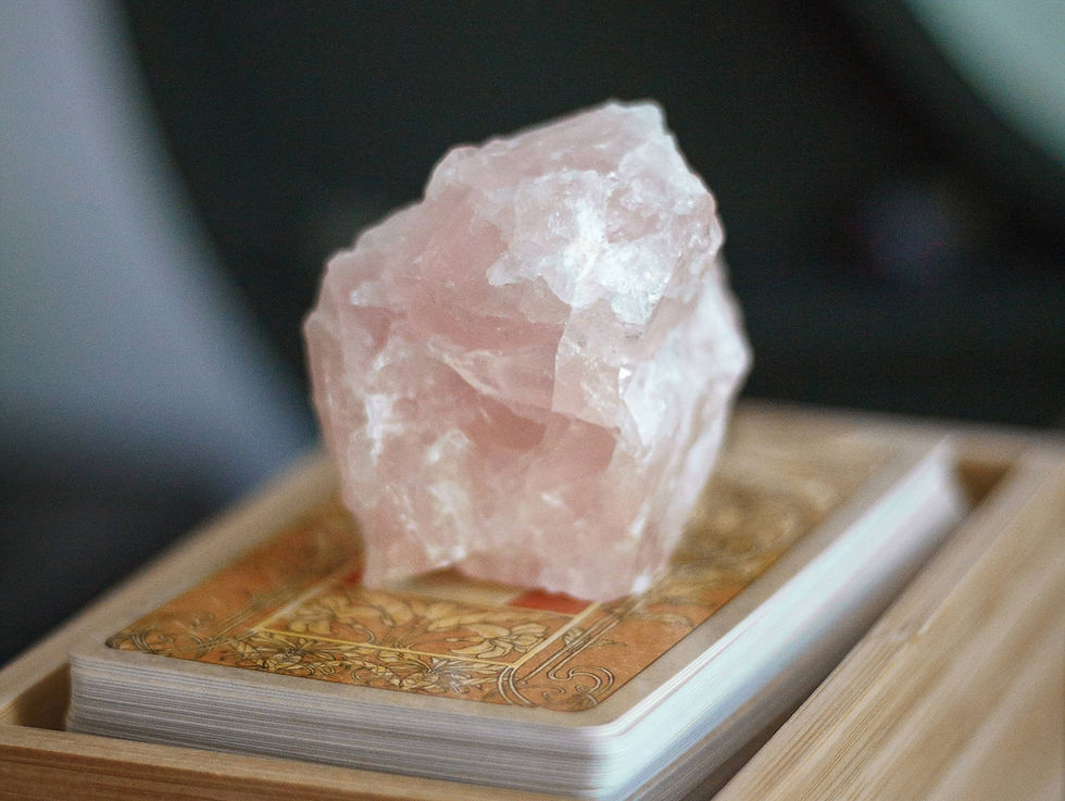 A rough pink crystal sits atop a stack of tarot cards with ornate designs. The setting is softly lit, creating a mystical ambiance.