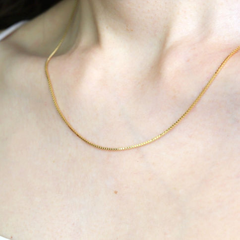 Close-up of a person's neck wearing a thin gold box chain on a skin. The mood is elegant and minimalistic.