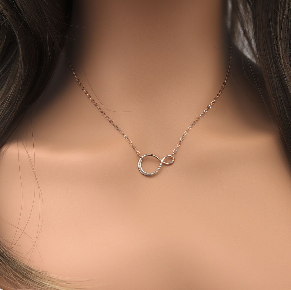 A rose gold infinity necklace on a woman's neck with brown hair. The background is softly blurred, elegant jewelry