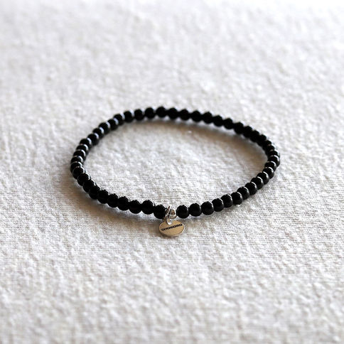 Black tourmaline beaded bracelet with a small metal tag on a textured white surface. The tag has a word engraved on it.