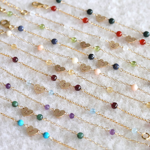 Gold chain bracelets with colorful beads and heart charms displaying zodiac signs are arranged on a textured white surface.