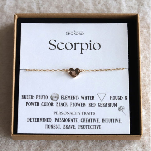 Gold heart anklet with Scorpio symbol on a card detailing zodiac traits and elements in a box. Text includes "SHOKORO".