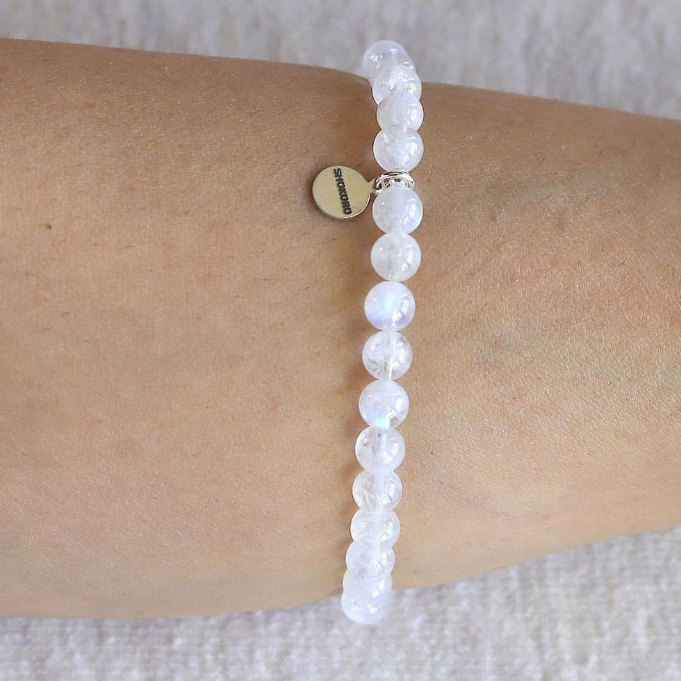 A moonstone beaded bracelet with translucent white beads and a small round tag is worn on a wrist. Background is a soft beige