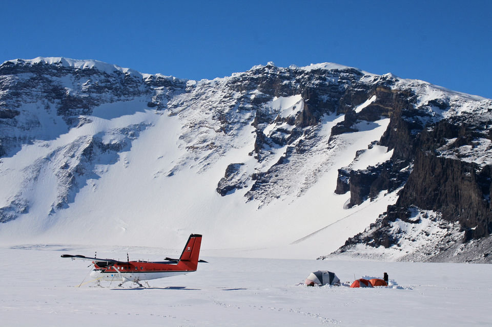 Two trekkers in blue jackets stand beside a large traditional Buddhist prayer wheel at what appears to be a monastery or temple, with mountains visible in the background, representing cultural immersion during mountain expeditions.