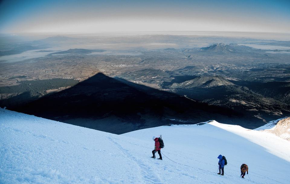 Aerial or high-altitude view showing climbers as small silhouettes on a snowy mountain ridge, with layers of mountains and valleys fading into the distance under a hazy sky at dawn or dusk.