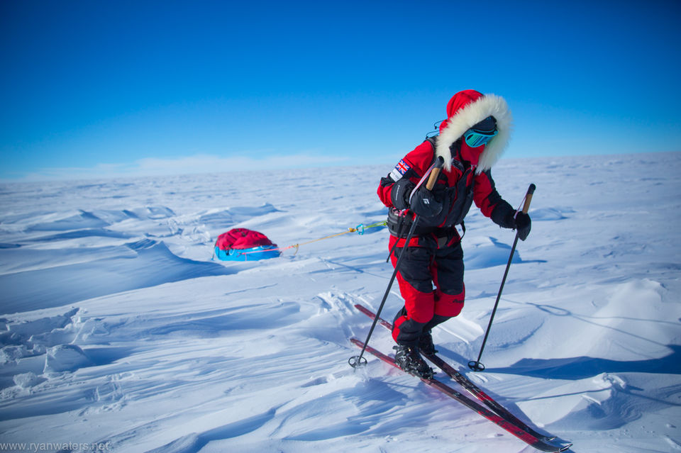 Close-up of expedition member in red polar suit and fur-trimmed hood skiing while pulling a blue sled, with snow formations visible in the background under bright blue skies.