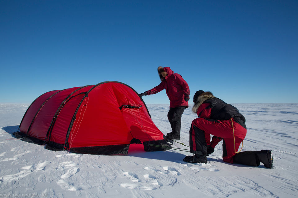 Person in red polar suit kneels beside a bright red dome tent pitched on pristine white snow under clear blue skies, setting up or organizing camp.