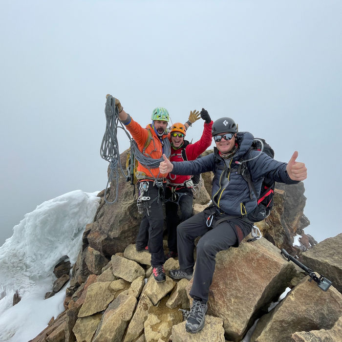 Climbers celebrate at a rocky summit, clustered around a summit marker or cairn, with misty mountain peaks visible in the background under overcast skies.