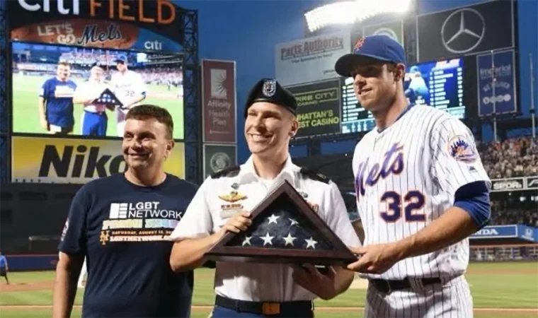 Josh Gravett, a soldier in uniform, standing on a baseball field with a member of the LGBT Network and a Mets player. They are handing a tri-folded American flag to Josh Gravett, symbolizing honor and service. The stadium audience is in the background.