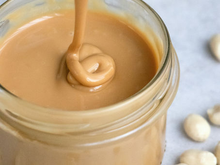 "Close-up of creamy peanut butter being spooned from a glass jar, surrounded by scattered peanuts, a visual cue for emotional eating or binge eating moments, especially tied to comforting foods like peanut butter."