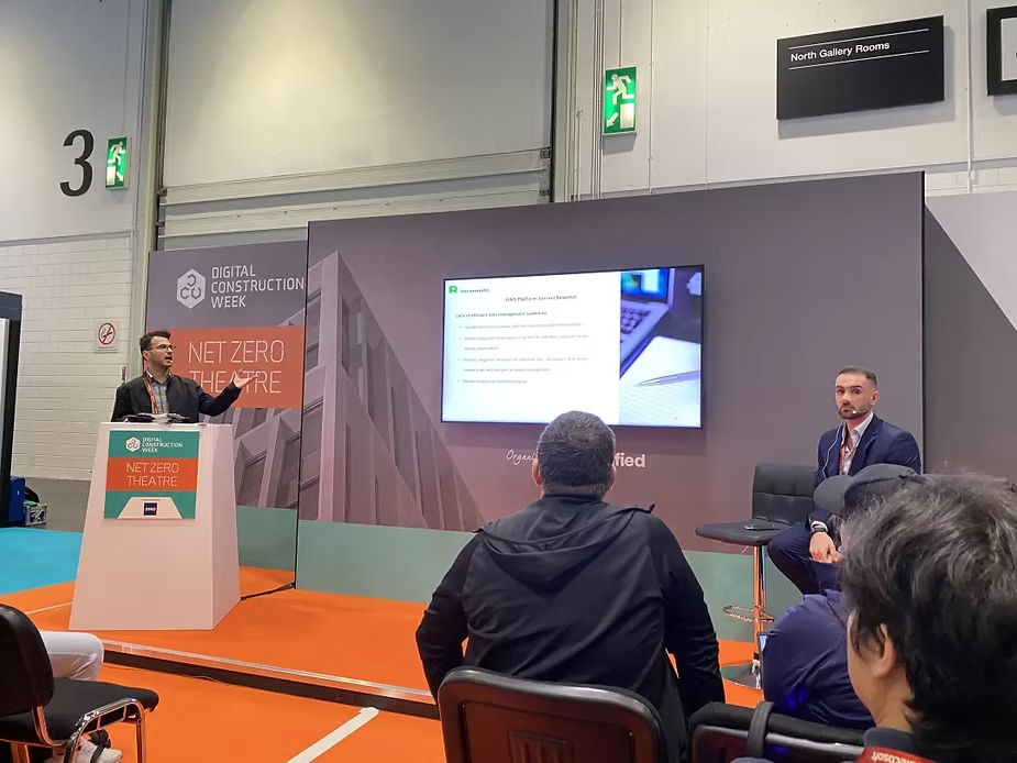 A speaker at a podium presents on "Digital Construction Week" in a conference room with "NET ZERO THEATRE" signage. Audience listens attentively.