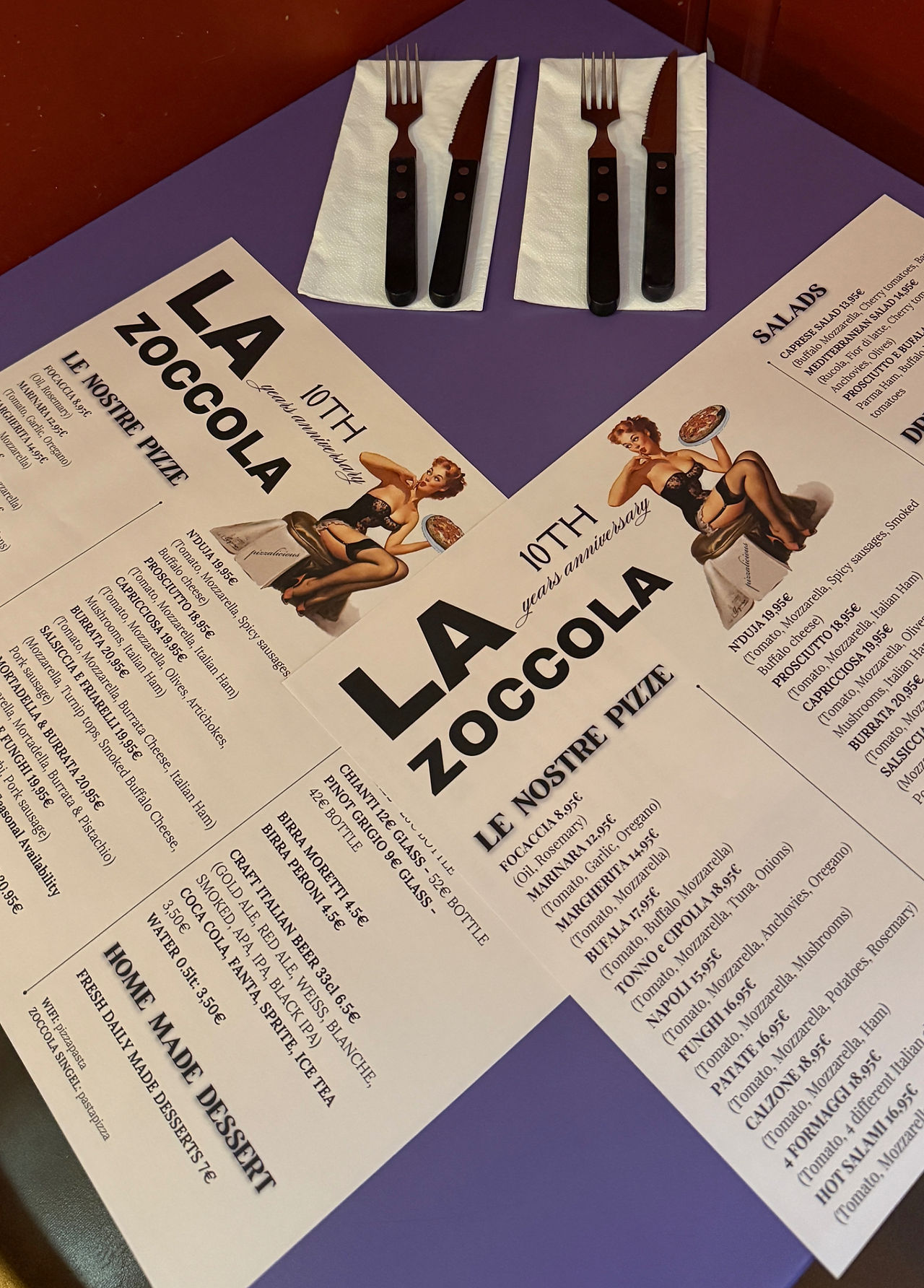 A high-angle shot of two paper menus on a purple table. The menu is for a restaurant named "LA ZOCCOLA." Two sets of black-handled cutlery on white napkins are placed next to the menus. The menus list different types of pizzas, salads, and drinks.