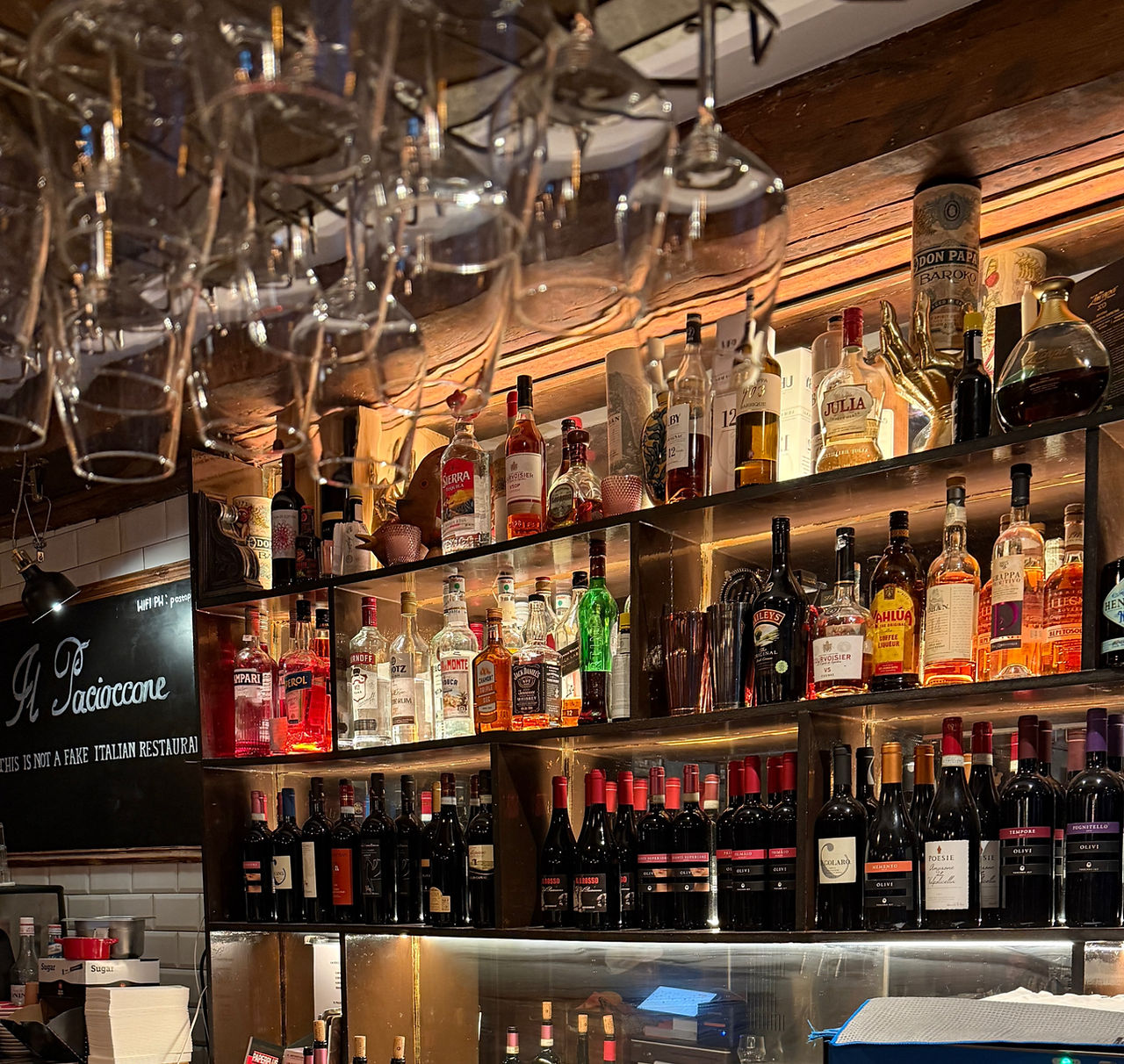 An indoor shot of a fully stocked bar. The bar counter is in the foreground, with a Marshall brand speaker. The background features several shelves lined with various bottles of wine and liquor. A row of wine glasses hangs upside down from a rack above the bar.