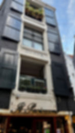 A low-angle shot of a tall, thin building in Amsterdam. The bottom of the building has a black sign with the name "Il Pacioccone" in gold cursive font. The dark facade is broken up by white window frames, with black wooden shutters on either side.