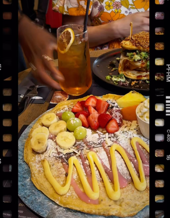 A flat-lay photo of a large, thin crêpe-style pancake on a blue speckled plate. The pancake is topped with sliced strawberries, banana rounds, green grapes, and a small wedge of orange and pineapple. A small paper cup of whipped cream sits near the top. The pancake is drizzled with a zigzag pattern of white and pink sauces, and dusted with powdered sugar and what appears to be grated chocolate. In the upper left corner, another plate with a stacked savory pancake is out of focus.
