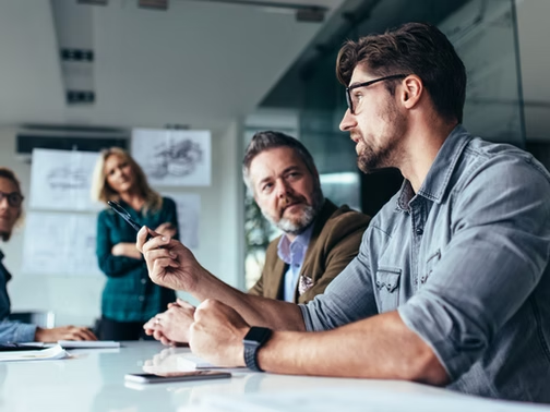 Man speaking with a pen at a conference table while coworkers listen in a modern office meeting room.