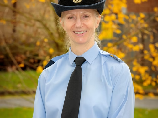 Smiling woman in a blue uniform shirt, black tie, and hat stands outdoors with autumn leaves in the background.