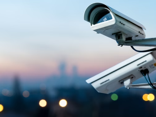 Two white security cameras on a pole overlook a blurred city skyline at dusk, with soft pink-blue sky and lights below.