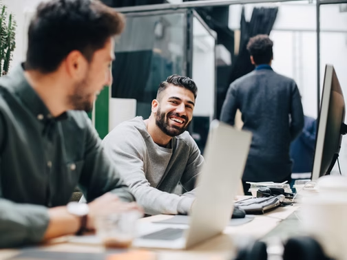 Smiling man at a laptop in a modern office, with coworkers nearby and one person walking away in the background.
