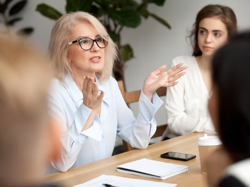 Older woman in glasses gestures while speaking at a meeting, with a younger woman listening at a table with notebook and phone.