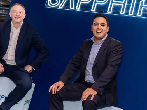 Two smiling men in suits sit on white cubes in a blue office, beneath a neon SAPPHIRE sign.