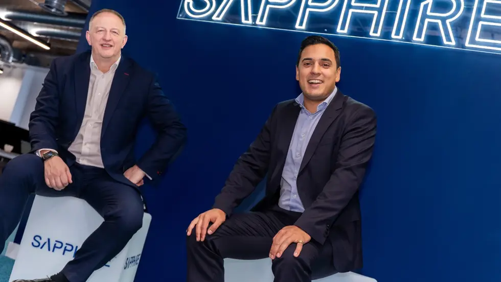 Two smiling men in suits sit on white cubes in a blue office, beneath a neon SAPPHIRE sign.