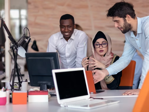 Three coworkers huddle around a laptop in a modern office, one man gesturing while others watch and smile.